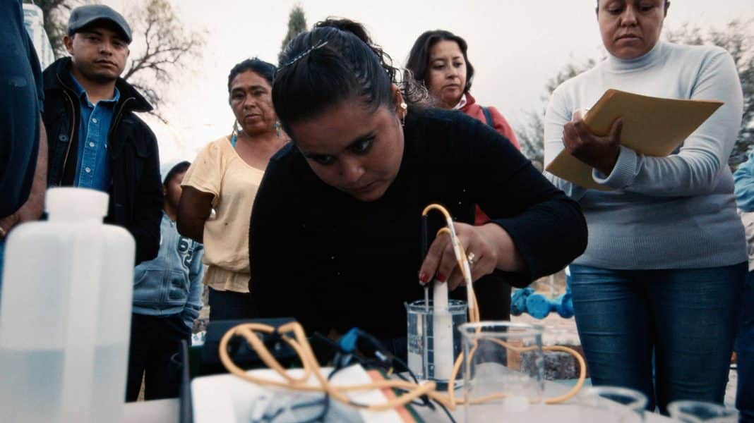 A small group of people gather outdoors around a table with scientific equipment. In the foreground, a Mexican woman in a black sweater is handling testing equipment and clear containers of liquid, while others observe. She wears a look of focus and determination of her face.
